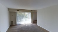 This single-floor residence offers a clean, functional layout designed for comfortable everyday living. The living area is finished with wall-to-wall carpeting that adds warmth underfoot, while neutral-toned walls provide a versatile backdrop for a variety of décor styles. A standout feature of the space is the expansive sliding glass door framed by vertical blinds, which allows generous amounts of natural light to pour into the room throughout the day. The large glazed opening also offers views of the surrounding outdoor greenery, creating a pleasant visual connection to the exterior. Ceiling-mounted HVAC vents are positioned strategically to ensure consistent climate control throughout the living area. The open flow between rooms allows for flexible furniture arrangements and a sense of spaciousness. Clean white baseboards and smooth ceilings contribute to the overall bright and tidy aesthetic of the interior. The understated, move-in-ready design of this space makes it an accessible and practical option for a range of occupants.