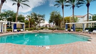 Located at 5010 Central Sarasota Pkwy in Sarasota, Florida, this resort-style outdoor amenity space delivers a lush, tropical retreat designed for relaxation and recreation. The centerpiece is a generously sized freeform swimming pool accompanied by a dedicated spa/hot tub, both finished with a brick paver surround that adds a refined, resort-quality aesthetic. Surrounding the pool deck are multiple private cabanas outfitted with white canopies and blue drapery, offering shaded lounging areas alongside rows of chaise lounge chairs accented with coordinating blue cushions and bright yellow umbrellas. A series of white pergolas are positioned throughout the grounds, providing additional shaded seating with outdoor furniture ideal for casual gathering. Meandering concrete pathways wind through meticulously maintained tropical landscaping, featuring mature live oaks, towering royal palms, sabal palms, and manicured hedgerows with decorative white rock accents. A central water feature with a fountain serves as a focal point along the main walkway, flanked by symmetrical pergola structures. The grounds are enclosed by ornamental black iron fencing, with views of the broader residential community visible in the background. The overall ambiance is bright, open, and inviting, reflecting the natural beauty characteristic of Southwest Florida's outdoor living.