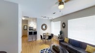 This single-floor residence showcases a thoughtfully designed open-concept layout that effortlessly connects the living, dining, and kitchen areas into one cohesive and functional living space. Warm hardwood flooring runs throughout, adding a sense of continuity and natural warmth to the interior. The living area is generously sized, accommodating a comfortable seating arrangement centered around a stylish coffee table, and is equipped with a ceiling fan that enhances airflow and comfort. Large windows fitted with white horizontal blinds allow abundant natural light to fill the space while maintaining privacy. The dining area sits centrally between the living space and kitchen, featuring a round table with modern chairs — an ideal setup for casual meals or entertaining. The kitchen is well-appointed with stainless steel appliances, including a microwave and range, complemented by a breakfast bar with bar-height seating and pendant lighting overhead. Track lighting adds a modern, functional touch to the kitchen zone. The walls are finished in a soft neutral gray tone, providing a clean and versatile backdrop throughout. The overall ambiance is bright, airy, and contemporary, with a practical yet stylish design that makes efficient use of the available space.