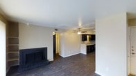 This single-story space in Garland offers an open-concept layout that seamlessly connects the living area to the kitchen. The living room features dark hardwood-style flooring that extends throughout, a brick fireplace with a dark surround serving as a focal point along one wall, and a ceiling fan that adds both functionality and a finishing touch to the space. Neutral white walls and white ceilings enhance the brightness of the room, creating a clean and airy atmosphere.

The kitchen is visible from the living area and is equipped with dark cabinetry that contrasts with the lighter walls, a decorative tile backsplash, and black appliances. A peninsula or partial wall separates the kitchen from the living space while maintaining an open feel between the two areas.

A hallway visible from the main living area leads to additional rooms beyond, suggesting a practical and well-organized floor plan. The consistent flooring and neutral color palette throughout create a cohesive aesthetic. Overall, the space combines functional design elements with a modern, understated style suited for comfortable everyday living.