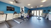 This single-floor fitness center is designed to support a comprehensive workout experience in a clean, motivating environment. The space is defined by bold blue accent walls paired with white wainscoting, creating a dynamic and energizing atmosphere throughout. Durable rubber flooring provides a safe, impact-absorbing surface across the entire facility.

The cardio section is well-appointed with multiple treadmills, elliptical trainers, and stationary bikes — including both upright and recumbent models — from brands such as Octane and True Fitness. A strength training area offers a multi-station cable machine, resistance bands, a stability ball, and a dedicated dumbbell rack with a water cooler nearby for convenience.

Large floor-to-ceiling mirrors line one wall, visually expanding the space and allowing users to monitor their form during workouts. Motivational athletic silhouette murals are featured prominently on the walls, adding an inspirational touch to the décor. Ceiling fans with flush-mount lighting fixtures provide comfortable air circulation and ample illumination.

A set of white-framed French doors floods the space with natural light and offers views of lush greenery and open outdoor surroundings, lending an airy, open feel. A wall-mounted flat-screen television adds an entertainment option for those engaged in longer cardio sessions. The overall design balances functionality with a welcoming aesthetic.