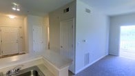 This single-floor residence offers a functional and straightforward layout designed for comfortable everyday living. The space is finished with neutral, light-toned walls throughout, creating a clean and bright atmosphere that feels open and welcoming.

The kitchen area features a raised bar-style counter with a light granite or laminate surface, providing a natural transition into the adjoining living space. A stainless steel sink is visible at the counter, indicating a practical and well-appointed kitchen setup. The flooring in the living area is carpeted in a neutral tone, adding warmth underfoot.

Ceiling-mounted HVAC vents are present throughout, ensuring consistent climate control across the space. Additional floor-level vents are also visible along the baseboards, supporting efficient airflow throughout the home.

Multiple six-panel white interior doors are visible, leading to separate rooms and offering privacy and clear spatial delineation. The doors and white trim provide a classic, tidy finish that complements the neutral color palette of the walls.

Overall, this space presents a clean, move-in-ready environment with practical amenities, a cohesive design aesthetic, and a layout that balances open communal areas with defined private spaces.