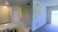 This single-floor residence offers a functional and straightforward layout designed for comfortable everyday living. The space is finished with neutral, light-toned walls throughout, creating a clean and bright atmosphere that feels open and welcoming.

The kitchen area features a raised bar-style counter with a light granite or laminate surface, providing a natural transition into the adjoining living space. A stainless steel sink is visible at the counter, indicating a practical and well-appointed kitchen setup. The flooring in the living area is carpeted in a neutral tone, adding warmth underfoot.

Ceiling-mounted HVAC vents are present throughout, ensuring consistent climate control across the space. Additional floor-level vents are also visible along the baseboards, supporting efficient airflow throughout the home.

Multiple six-panel white interior doors are visible, leading to separate rooms and offering privacy and clear spatial delineation. The doors and white trim provide a classic, tidy finish that complements the neutral color palette of the walls.

Overall, this space presents a clean, move-in-ready environment with practical amenities, a cohesive design aesthetic, and a layout that balances open communal areas with defined private spaces.