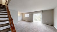 This two-story home offers a functional and straightforward layout spread across two floors. The main level houses the living room and kitchen, while the upper floor contains the two bedrooms, bathroom, hallway, and ample closet storage across four closets.

The living room is a generous, open space finished with plush wall-to-wall carpeting in a neutral gray tone. Large vertical-blind-covered sliding glass doors flood the room with natural light and provide views of surrounding greenery, while an additional window further brightens the space. A wood-railed staircase connects both floors, adding a warm accent to the interior.

The kitchen features white cabinetry, a speckled light countertop, a stainless steel sink, and a gas range, offering a compact and efficient cooking space with tile flooring.

Upstairs, the primary bedroom is a comfortable, well-sized room with carpet flooring and a window allowing in natural light. The second bedroom is similarly carpeted and bright. Both rooms are served by a shared full bathroom, which includes a vanity with a white cabinet, a mirror, a toilet, a shower/tub combination, and wood-look flooring. Wall sconce lighting adds a clean, classic touch throughout the upper level.

Overall, the home presents a clean, neutral palette with practical finishes throughout.