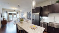 This thoughtfully designed single-floor residence showcases a seamless open-concept layout that effortlessly connects the kitchen and living areas. The kitchen is a standout feature, anchored by a generous granite-topped island with seating — ideal for casual dining or entertaining. Dark espresso cabinetry provides a striking contrast against the light stone countertops, while stainless steel appliances, including a French door refrigerator with ice dispenser and a dishwasher, offer a sleek, modern aesthetic. Elegant glass pendant lights hang above the island, adding a refined touch to the space. A classic subway tile backsplash completes the kitchen's polished look. The open living area beyond is bright and welcoming, benefiting from natural light that filters through large windows. A ceiling fan enhances comfort, while the neutral color palette and contemporary furnishings create a warm, inviting ambiance. Curated wall art adds personality to the space. The overall design strikes a balance between modern sophistication and everyday livability, making this a highly functional and visually appealing home.