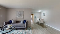 This single-floor unit offers a thoughtfully designed layout with a seamless flow between the main living spaces. The combined living and dining area is finished with light wood-tone luxury vinyl flooring, creating a bright and cohesive atmosphere. Neutral warm-gray walls provide a versatile backdrop throughout.

The living area is well-proportioned, anchored by a tufted gray sofa, a glass-top coffee table, and a vibrant teal-accented area rug. A large sliding glass door floods the space with natural light and provides access to an outdoor balcony with exterior views. The dining area, positioned adjacent to the living room, accommodates a white dining table and chairs and sits near a conveniently located stacked washer and dryer unit, offering practical in-unit laundry.

The bedroom features soft carpeting, a ceiling fan, a baseboard heating unit, and a window dressed with blinds that allows natural light to filter in comfortably.

The bathroom is finished with large-format light tile flooring, white subway tile wainscoting, a white vanity, and a shower-tub combination, presenting a clean and functional design. Overall, the space maintains a modern, neutral aesthetic throughout.