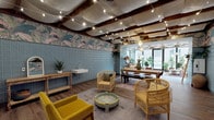 This single-floor space showcases a richly layered bohemian aesthetic unified by a consistent design language throughout. The ceiling is a defining architectural feature, with exposed dark wood beams supporting billowing white fabric panels and strung with warm globe lights that create an intimate, canopy-like atmosphere overhead.

Walls are adorned with bold tropical botanical wallpaper featuring lush palm and bird motifs in earthy pinks, greens, and blues, offset by lower sections clad in muted blue square tiles. These elements combine to give the interior a distinctive, eclectic character. The flooring transitions between warm wood-tone planks and light wood-effect surfaces, grounding the vibrant décor.

The main lounge area is furnished with rattan armchairs, a mustard yellow sofa, a painted tray-style coffee table, and layered vintage rugs, creating an inviting, relaxed seating arrangement. A rustic wood console with a wall-mounted sink adds a functional yet stylish element to this zone.

A sun-filled glazed room anchors one end of the space, with floor-to-ceiling windows on multiple sides drawing in abundant natural light and outdoor views. Lush potted plants, rattan seating, and patterned cushions enhance the tropical, garden-room feel. A dining area with a long wooden table bridges the two zones, completing this generous and visually rich space.