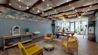 This single-floor space showcases a richly layered bohemian aesthetic unified by a consistent design language throughout. The ceiling is a defining architectural feature, with exposed dark wood beams supporting billowing white fabric panels and strung with warm globe lights that create an intimate, canopy-like atmosphere overhead.

Walls are adorned with bold tropical botanical wallpaper featuring lush palm and bird motifs in earthy pinks, greens, and blues, offset by lower sections clad in muted blue square tiles. These elements combine to give the interior a distinctive, eclectic character. The flooring transitions between warm wood-tone planks and light wood-effect surfaces, grounding the vibrant décor.

The main lounge area is furnished with rattan armchairs, a mustard yellow sofa, a painted tray-style coffee table, and layered vintage rugs, creating an inviting, relaxed seating arrangement. A rustic wood console with a wall-mounted sink adds a functional yet stylish element to this zone.

A sun-filled glazed room anchors one end of the space, with floor-to-ceiling windows on multiple sides drawing in abundant natural light and outdoor views. Lush potted plants, rattan seating, and patterned cushions enhance the tropical, garden-room feel. A dining area with a long wooden table bridges the two zones, completing this generous and visually rich space.