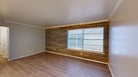 This single-floor unit combines rustic character with clean, modern finishes throughout its well-organized layout. The most distinctive feature is the expansive reclaimed wood accent wall in the main living area, where horizontal planks in varying earth tones create a warm, textured focal point. Crown molding frames the ceiling, and a large window fitted with horizontal blinds allows natural light to fill the space. Wood-look flooring runs consistently through the living area, complementing the accent wall's warm palette.

The entryway is straightforward and functional, with flush-mounted ceiling lighting and light-toned wood-look flooring that transitions into the adjoining rooms. A secondary room features neutral gray walls, a solid white door, and the same durable flooring, offering a versatile space suitable for a variety of uses.

The kitchen is efficiently arranged, featuring gray flat-front cabinetry with brushed metal hardware, white countertops, and a full-size refrigerator. Light-toned flooring and a door leading to an adjacent area keep the space practical and connected.

The bathroom is clean and bright, with a full tub-shower combination framed by white tile surround, a curved shower rod, a white vanity with gray cabinetry below, and light wood-look flooring. The overall ambiance of the unit is neutral and move-in ready.