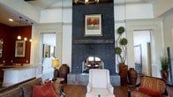 This single-floor space makes a striking first impression with its dramatic, floor-to-ceiling fireplace clad in dark, textured stone that serves as the undeniable focal point of the main living area. The fireplace surround extends to impressive heights, crowned by a rustic wooden mantel and a framed landscape painting that adds a touch of classic elegance. An arched pass-through at the base of the fireplace offers a unique architectural detail, providing a glimpse into an adjoining space beyond.

The soaring ceilings lend an open, grand sense of scale to the interior, enhanced by clean white walls adorned with detailed molding and trim work. Warm hardwood flooring grounds the space, complementing the rich tones of the furnishings, including upholstered armchairs, classic side tables, and decorative ceramic urns. Lush topiary-style plants flank the fireplace, adding organic texture and life to the setting.

Natural light filters in from surrounding windows and a visible adjoining room with large glass elements, keeping the atmosphere bright and airy. The overall ambiance is refined yet comfortable, blending traditional architectural details with warm, inviting décor that creates a sophisticated and welcoming environment throughout.