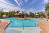 Located at 8160 Veterans Pkwy in Columbus, Georgia, this outdoor amenity complex offers a thoughtfully designed aquatic retreat for residents. The centerpiece of the space is a generously sized, freeform swimming pool with a smooth concrete surround, lined with blue tile accents and equipped with entry handrails for ease of access. Surrounding the pool, a broad deck accommodates an array of lounge chairs and shaded umbrella tables, ideal for relaxing in the Georgia sunshine.

A second, shallower pool area serves as a dedicated splash pad and wading zone, complete with interactive water features including spray elements — making it a family-friendly destination. The brick-paver patio extends the usable outdoor space, providing additional seating areas framed by lush, manicured landscaping.

The grounds are bordered by a striking natural stone retaining wall, rising against a backdrop of mature evergreen and deciduous trees that lend privacy and a park-like ambiance. A covered pavilion with an attached structure provides shaded gathering space adjacent to the pools. The overall design harmoniously blends functional aquatic amenities with attractive green spaces, creating an inviting outdoor environment well-suited to both active recreation and leisurely relaxation.