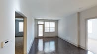 This single-floor space presents a clean, modern aesthetic defined by its polished dark hardwood flooring, which reflects natural light and adds a sense of depth to the interior. The main living area is characterized by crisp white ceilings and light gray walls, creating a neutral and versatile backdrop. A combination of a glass-paneled door and an adjacent large window with horizontal blinds dominates one wall, allowing generous amounts of natural light to fill the room while offering access to what appears to be a balcony or exterior area. The flooring's high-gloss finish enhances the brightness of the space, complementing the clean lines throughout. White baseboards provide a crisp contrast against the dark floors, contributing to the polished overall appearance. Floor-level HVAC vents are integrated along the walls, maintaining the streamlined aesthetic. An arched doorway on one side leads to an adjacent area, suggesting a practical, open layout. The space's minimal design language and quality finishes make it an adaptable and well-appointed environment.