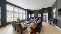 Located at 5400 Chenonceau Blvd in Little Rock, Arkansas, this single-floor community space offers a sophisticated and well-appointed gathering area designed to serve a variety of needs. The interior is characterized by deep charcoal-toned walls accented with crisp white trim and molding panels, creating a bold yet refined aesthetic throughout.

The main lounge area is generously sized, featuring light-toned tile flooring and a comfortable arrangement of upholstered sofas and accent chairs layered over a patterned area rug. Decorative throw pillows in vibrant patterns add visual interest, while sculptural side tables and curated wall art contribute to the space's polished character. A wall-mounted flat-screen television anchors one side of the lounge, making it suitable for casual viewing or social gatherings.

Tall windows along one wall flood the space with abundant natural light, offering views of the surrounding greenery and creating an open, airy atmosphere despite the darker wall palette. Adjacent to the lounge, a café-style seating area with bar-height tables and stools provides an informal spot for conversation or individual work.

A dedicated business center is seamlessly integrated into the space, featuring a long countertop workstation equipped with desktop computers and a printer, illuminated by striking geometric pendant light fixtures. The overall ambiance is contemporary and inviting, blending functional amenities with thoughtful design details throughout.