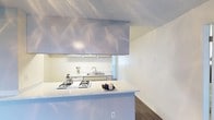 This single-floor space offers a clean, minimalist aesthetic defined by bright white walls, dark hardwood-style flooring, and a textured ceiling that adds subtle visual character throughout. The kitchen serves as a central feature, designed around an open peninsula layout with a light-toned stone-finish countertop that provides generous prep space and a natural gathering point. A built-in gas cooktop sits flush within the peninsula surface, while a large overhead range hood anchors the kitchen visually and provides practical ventilation. Beyond the peninsula, a secondary counter runs along the wall and houses a stainless steel sink positioned beneath a pass-through window, enhancing the sense of openness and allowing natural light to filter into the cooking area. The kitchen cabinetry and surfaces are finished in a consistent soft white palette, contributing to a cohesive and airy feel. The transition between the kitchen and adjacent living areas is seamless, with the dark flooring providing a grounding contrast to the lighter walls and countertops. The overall ambiance of the space is light, clean, and functional — well-suited for everyday living with an emphasis on simplicity and practicality.