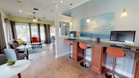 This single-story community space is designed to offer both functionality and comfort, seamlessly blending several distinct areas into a cohesive, welcoming environment. The main lounge area is anchored by large windows dressed with floor-length gray drape panels, allowing abundant natural light while framing picturesque views of an outdoor pool and a tranquil waterway lined with palm trees. A tray ceiling with a ceiling fan adds architectural definition to the seating area, which features a tufted sofa, a tile-surround fireplace, and decorative round mirrors on the wall.

Adjacent to the lounge is a casual seating nook with a loveseat and bistro-style table, also enjoying direct views of the pool area through sliding glass doors. The kitchen and bar area is well-equipped with warm wood cabinetry, a mosaic tile backsplash, granite-style countertops, pendant lighting, and a peninsula with bar seating — ideal for casual gatherings. A separate business or workstation bar counter, accented by pendant lights and a large coastal-themed painting, provides a functional workspace. Throughout, terra cotta tile flooring ties the areas together, contributing to the bright, resort-inspired ambiance of the overall space.