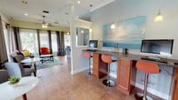 This single-story community space is designed to offer both functionality and comfort, seamlessly blending several distinct areas into a cohesive, welcoming environment. The main lounge area is anchored by large windows dressed with floor-length gray drape panels, allowing abundant natural light while framing picturesque views of an outdoor pool and a tranquil waterway lined with palm trees. A tray ceiling with a ceiling fan adds architectural definition to the seating area, which features a tufted sofa, a tile-surround fireplace, and decorative round mirrors on the wall.

Adjacent to the lounge is a casual seating nook with a loveseat and bistro-style table, also enjoying direct views of the pool area through sliding glass doors. The kitchen and bar area is well-equipped with warm wood cabinetry, a mosaic tile backsplash, granite-style countertops, pendant lighting, and a peninsula with bar seating — ideal for casual gatherings. A separate business or workstation bar counter, accented by pendant lights and a large coastal-themed painting, provides a functional workspace. Throughout, terra cotta tile flooring ties the areas together, contributing to the bright, resort-inspired ambiance of the overall space.