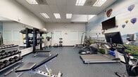 This fitness center is a compact yet efficiently organized workout space designed to accommodate a wide range of exercise needs. The room features durable rubber flooring in a speckled blue-grey finish, providing both safety and comfort underfoot. Overhead, a drop ceiling with recessed fluorescent panel lighting ensures the space is consistently and evenly illuminated throughout.

The cardio section is anchored by TRUE-brand treadmills, an elliptical trainer, and an Echelon rowing machine, offering diverse options for aerobic conditioning. Strength training is well-supported with a Hoist multi-station cable machine, adjustable weight benches, a barbell rack with plate weights, and a full dumbbell rack. Additional functional training accessories include stability balls and resistance bands, rounding out the facility's versatility.

A large, eye-catching wall mural depicting a colorful hot air balloon festival over a scenic landscape serves as a standout design feature, adding energy and visual interest to the space. Full-length mirrors along one wall enhance the sense of openness and allow users to monitor their form during workouts. Frosted glass panels and a glass-paned exit door allow natural light to filter in while maintaining privacy. A wall clock and signage complete the practical, user-focused environment.