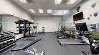 This fitness center is a compact yet efficiently organized workout space designed to accommodate a wide range of exercise needs. The room features durable rubber flooring in a speckled blue-grey finish, providing both safety and comfort underfoot. Overhead, a drop ceiling with recessed fluorescent panel lighting ensures the space is consistently and evenly illuminated throughout.

The cardio section is anchored by TRUE-brand treadmills, an elliptical trainer, and an Echelon rowing machine, offering diverse options for aerobic conditioning. Strength training is well-supported with a Hoist multi-station cable machine, adjustable weight benches, a barbell rack with plate weights, and a full dumbbell rack. Additional functional training accessories include stability balls and resistance bands, rounding out the facility's versatility.

A large, eye-catching wall mural depicting a colorful hot air balloon festival over a scenic landscape serves as a standout design feature, adding energy and visual interest to the space. Full-length mirrors along one wall enhance the sense of openness and allow users to monitor their form during workouts. Frosted glass panels and a glass-paned exit door allow natural light to filter in while maintaining privacy. A wall clock and signage complete the practical, user-focused environment.