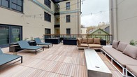 This outdoor space is composed of two distinct deck areas finished with warm wood decking throughout. The upper terrace is an open-air lounge designed for gathering, featuring modern white-frame sectional sofas dressed in tan cushions arranged around a rectangular fire table. Black raised planters add greenery, and overhead string lights create an inviting ambiance. A wood-and-iron railing frames the perimeter, offering open views of the surrounding urban neighborhood and neighboring rooftops.

The lower courtyard-level deck is nestled between the building's brick and smooth stucco exterior walls, creating a more sheltered atmosphere. This area is furnished with sleek lounge chairs and additional raised planters filled with ornamental grasses and white stone. A glass-enclosed corridor connects to the building's interior, allowing natural light to filter through. Decorative tile detailing along the building's base adds a thoughtful architectural accent. String lights are strung across both levels, lending a cohesive and welcoming character to the entire outdoor environment, day or night.