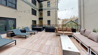 This outdoor space is composed of two distinct deck areas finished with warm wood decking throughout. The upper terrace is an open-air lounge designed for gathering, featuring modern white-frame sectional sofas dressed in tan cushions arranged around a rectangular fire table. Black raised planters add greenery, and overhead string lights create an inviting ambiance. A wood-and-iron railing frames the perimeter, offering open views of the surrounding urban neighborhood and neighboring rooftops.

The lower courtyard-level deck is nestled between the building's brick and smooth stucco exterior walls, creating a more sheltered atmosphere. This area is furnished with sleek lounge chairs and additional raised planters filled with ornamental grasses and white stone. A glass-enclosed corridor connects to the building's interior, allowing natural light to filter through. Decorative tile detailing along the building's base adds a thoughtful architectural accent. String lights are strung across both levels, lending a cohesive and welcoming character to the entire outdoor environment, day or night.