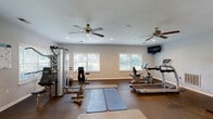 Located at 1225 Boone Hill Rd in Summerville, SC, this dedicated exercise room offers a well-appointed fitness facility designed to support a comprehensive workout routine. The space is housed on a single floor and provides a generous open layout with durable rubber flooring suited for high-impact activity.

The room is outfitted with multiple cardio machines, including a treadmill, stationary bike, and elliptical trainer, alongside a multi-station strength training machine. Exercise mats and a stability ball round out the functional fitness offerings. A wall-mounted multi-cable system adds further versatility for strength and conditioning exercises.

Recessed lighting illuminates the space evenly throughout, while two ceiling fans promote comfortable air circulation during workouts. Multiple wall-mounted flat-screen TVs are positioned at convenient viewing angles, enhancing the exercise experience. Natural light filters in through several windows fitted with white blinds, providing views of the surrounding greenery and keeping the environment bright and inviting.

The interior features clean, neutral-toned walls with white trim and baseboards, lending a crisp, modern aesthetic. A dedicated entrance door with an emergency exit sign and a fire extinguisher reflect safety-conscious design. The overall ambiance is practical, clean, and well-maintained, making this fitness room a fully equipped space for residents seeking convenient on-site exercise amenities.
