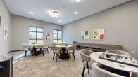 Located at 6620 Chestnut St in Lino Lakes, Minnesota, this dedicated game room offers a well-designed communal space built entirely around leisure and social interaction. The room is finished with neutral gray walls and patterned carpet flooring that lends a modern, welcoming aesthetic throughout.

The space is furnished with multiple round and rectangular tables paired with curved, upholstered chairs in warm beige and gray tones, providing ample seating for groups. A standout feature is the marble-topped chess table, which serves as a central gathering point. A full-length shuffleboard table lines one wall, complemented by a classic arcade cabinet and a wall-mounted flat-screen television, offering a diverse range of entertainment options.

Natural light flows in through several large windows along the exterior wall, offering views of the surrounding outdoor greenery and keeping the space bright and airy. A cluster pendant light fixture and recessed ceiling lights provide additional illumination throughout the room.

Colorful, game-themed artwork — depicting playing cards, chess pieces, and board game motifs — adorns the walls, reinforcing the playful character of the space. Interior windows connect this room to adjacent areas, adding a sense of openness. Overall, the game room is thoughtfully curated to encourage community engagement and recreational activity.