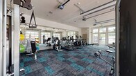 Located at 1161 3rd Ave E in Bradenton, FL, this fitness center offers a well-equipped, single-floor workout facility designed to support a full range of exercise needs. The space is generously proportioned, providing ample room for members to move freely between equipment zones without feeling crowded.

The flooring throughout features a distinctive teal and charcoal modular carpet tile pattern that adds a modern, energetic aesthetic to the space. Large glass garage-style doors and floor-to-ceiling windows line the walls, flooding the interior with abundant natural light and offering views of the surrounding outdoor environment. Full-length mirrors are strategically placed along the walls, enhancing the sense of space and allowing users to monitor their form.

The cardio zone is well-stocked with multiple treadmills, elliptical trainers, and stationary bikes, all arranged to face the mirrored walls. The strength training area includes a multi-station cable machine, a functional trainer with rope and strap attachments, a full dumbbell rack with an adjustable bench, and additional selectorized weight machines. A rowing machine rounds out the cardiovascular offerings.

The facility is bright, clean, and professionally maintained, with a high ceiling that contributes to an open, airy atmosphere. Multiple entry and exit points, including a dedicated side door, provide easy access throughout the space.