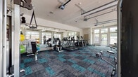 Located at 1161 3rd Ave E in Bradenton, FL, this fitness center offers a well-equipped, single-floor workout facility designed to support a full range of exercise needs. The space is generously proportioned, providing ample room for members to move freely between equipment zones without feeling crowded.

The flooring throughout features a distinctive teal and charcoal modular carpet tile pattern that adds a modern, energetic aesthetic to the space. Large glass garage-style doors and floor-to-ceiling windows line the walls, flooding the interior with abundant natural light and offering views of the surrounding outdoor environment. Full-length mirrors are strategically placed along the walls, enhancing the sense of space and allowing users to monitor their form.

The cardio zone is well-stocked with multiple treadmills, elliptical trainers, and stationary bikes, all arranged to face the mirrored walls. The strength training area includes a multi-station cable machine, a functional trainer with rope and strap attachments, a full dumbbell rack with an adjustable bench, and additional selectorized weight machines. A rowing machine rounds out the cardiovascular offerings.

The facility is bright, clean, and professionally maintained, with a high ceiling that contributes to an open, airy atmosphere. Multiple entry and exit points, including a dedicated side door, provide easy access throughout the space.
