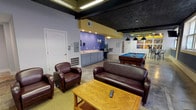 This single-floor community lounge is thoughtfully organized into several functional zones, offering a well-rounded social environment. The space is defined by its bold color palette — deep blue and vibrant purple accent walls contrast against light gray and white surfaces — complemented by a striking dark embossed tin ceiling that adds architectural character throughout.

The main seating area features rich burgundy leather sofas and armchairs arranged around a parquet-top wooden coffee table, creating a relaxed gathering spot. Large gridded windows along the blue-accented wall flood this area with natural light. A large projection wall nearby adds a media-viewing capability to the lounge.

Adjacent to the seating zone, the recreation area centers on a full-size pool table with a blue felt surface, suspended overhead by classic pendant lights. Wall-mounted televisions provide additional entertainment options throughout the space.

A kitchenette with gray cabinetry and a sink, set against a purple accent wall, offers convenient refreshment access. The open floor plan, featuring polished concrete flooring in areas of the space, transitions seamlessly between zones. Additional amenities include a water fountain, restroom access, a vending machine, and ample open floor space, making this lounge a versatile and inviting communal destination.