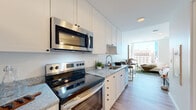 This contemporary single-floor residence showcases a thoughtfully designed open layout that maximizes both functionality and natural light. The galley-style kitchen is outfitted with a full suite of stainless steel Frigidaire appliances, including a French door refrigerator with ice dispenser, an electric range with over-the-range microwave, and a double basin sink. Crisp white shaker cabinetry extends to the ceiling, providing generous storage, while light grey granite countertops add a polished, refined finish. Light wood-tone flooring runs continuously throughout the space, reinforcing a clean and cohesive aesthetic.

The open living area flows seamlessly from the kitchen and is defined by dramatic floor-to-ceiling windows that frame sweeping views of the surrounding urban skyline. The abundance of natural light creates a bright, airy atmosphere throughout the day. A cozy reading nook positioned directly at the corner window offers an ideal vantage point over the cityscape below.

The bathroom features warm wood-toned cabinetry with a white stone countertop, an arched black-framed mirror, and light hardwood-style flooring, delivering a contemporary and well-appointed finish that complements the overall design sensibility of the space.