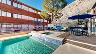 This outdoor amenity area is defined by a generously sized rectangular swimming pool with clear turquoise water, flanked by a concrete and paver deck outfitted with colorful lounge chairs and seating areas. A raised spa with blue tile accents adjoins the main pool, featuring a water cascade feature that flows into the pool below. The pool deck is enclosed by a black metal fence, providing both safety and a defined boundary for the space.

The surrounding multi-story brick building wraps around portions of the area, creating a semi-enclosed courtyard feel that offers a degree of shelter and privacy. Large windows on the building façade overlook the pool deck. Shaded seating areas equipped with blue patio umbrellas and bistro-style tables and chairs are positioned throughout, providing comfortable spots for relaxation outside the water.

A striking natural backdrop distinguishes this space: rugged rocky cliffs rise dramatically behind the complex, accompanied by a tall, mature pine tree that adds greenery and natural texture. The combination of the warm brick architecture, the natural stone escarpment, and the vibrant blue pool water creates a visually compelling outdoor environment ideal for recreation and leisure under open skies.