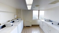 This shared laundry facility is a well-organized, single-floor space designed to accommodate multiple users efficiently. The room is outfitted with a full lineup of top-load washing machines and dryers arranged along two walls, providing ample capacity for simultaneous use. The flooring consists of light-toned ceramic tiles with blue grout lines, offering a clean and durable surface well-suited for a high-traffic utility space. Overhead, recessed fluorescent panel lighting ensures bright, even illumination throughout the room. A large window on the far wall allows natural light to fill the space while offering an elevated view of the surrounding tree canopy and cityscape below, adding a surprisingly pleasant visual element to an otherwise functional environment. The walls are finished in a neutral off-white, contributing to the room's clean and practical aesthetic. A wall-mounted utility box and shelving bracket are also present, providing additional functional support. Ventilation is handled by a ceiling-mounted exhaust unit, keeping the space fresh during operation. Overall, the laundry room is practical, well-lit, and efficiently laid out, making it a convenient and functional shared amenity.