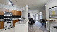 This single-floor residence offers a thoughtfully designed open-concept layout that seamlessly connects the kitchen and living areas, creating a spacious and inviting atmosphere. The kitchen is a standout feature, boasting granite countertops with subtle veining, warm honey-toned wood cabinetry, and a sleek black tile backsplash that adds a bold contrast. A peninsula with bar seating extends into the living space, providing both additional prep space and a casual dining option. Stainless steel appliances and a double basin sink complete the functional kitchen setup. An elegant decorative column with crown molding detailing serves as a graceful architectural divider between the kitchen and living room, adding a classic touch to the space. The living area is bright and airy, benefiting from multiple large windows dressed with plantation-style blinds that allow natural light to flood the room. A glass-paneled door offers direct access to the outdoors, further enhancing the sense of openness. Crown molding runs throughout the ceiling, lending a refined, polished character to the entire space. Neutral tones in the carpeting and walls create a versatile backdrop, making the residence feel both warm and welcoming.