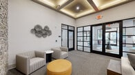 Located at 5000 Collina Ter in Clermont, FL, this thoughtfully designed living room delivers a polished, contemporary aesthetic across its single-floor layout. The space immediately impresses with its elevated coffered ceiling detailed with warm ambient LED uplighting, lending depth and sophistication to the room's architecture. Large steel-framed glass partitions and doors line one wall, flooding the interior with natural light while providing a seamless visual connection to adjacent areas beyond. The flooring throughout is a neutral, textured carpet that grounds the space with warmth and comfort. A striking stacked stone accent wall serves as a bold focal point, flanking a wall-mounted flat-screen display and complementing the earthy terracotta-toned wall panel between the stone columns. A generous sectional sofa anchors the main seating area, paired with a low-profile wooden coffee table and a live-edge wood console desk. Near the glass entry, a relaxed lounge nook features two upholstered accent chairs, a round white side table, and a vibrant yellow ottoman that adds a pop of color. Abstract wall art and sculptural metalwork wall décor contribute an artistic sensibility throughout, completing this refined and inviting living environment.