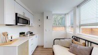 This single-floor studio is thoughtfully designed to maximize functionality within a compact footprint. The interior is finished with a clean, modern aesthetic, featuring crisp white walls, light wood-tone flooring, and white cabinetry throughout. The sleeping area is positioned near a corner of large windows fitted with horizontal blinds, allowing ample natural light to fill the space while offering views of the surrounding neighborhood. The multi-pane window configuration wraps around the corner, creating a bright, airy atmosphere.

The kitchenette is efficiently outfitted with white cabinetry above and below a light-toned countertop, a built-in stainless steel microwave, and a small sink — providing essential cooking and food preparation capabilities. Adjacent to the kitchenette, an open closet with built-in shelving offers organized storage.

The bathroom is well-appointed with a white vanity, neutral countertop, and a walk-in shower featuring marble-look tile walls and a classic striped curtain. A narrow window above the toilet brings in natural light. Wood-look flooring extends throughout, tying the spaces together with a cohesive, contemporary feel.