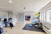Located at 19255 NE Third Ave in Miami, FL, this amenity space is divided into two functional areas designed to support both active fitness and leisure activities. The fitness area is generously appointed with a wide range of equipment, including treadmills, stationary and recumbent bicycles, a multi-station cable machine, a weight bench, a dumbbell rack, resistance bands, medicine balls, stability balls, and foam rollers organized on dedicated storage racks. A padded exercise mat anchors the floor space for stretching or functional training. A wall-mounted flat-screen TV adds convenience for workout sessions. Windows along the perimeter allow natural light to fill the room, complementing the recessed lighting in the drop tile ceiling. The light wood-style flooring runs throughout, giving the space a clean, cohesive look. Soft gray walls create a calm, neutral backdrop across both areas. The adjacent recreation room centers around a full-size pool table with a bold red felt surface, surrounded by classic wooden arm chairs for spectators. A small seating nook with blue leather chairs and a stone-top side table offers a relaxed lounge corner. Bookshelves line one wall, adding a casual, communal feel to the space.