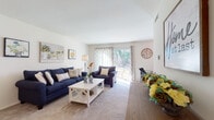 This single-floor residence offers a thoughtfully arranged layout that balances living, working, and resting spaces. The living room serves as the heart of the home, featuring plush wall-to-wall carpeting, neutral-toned walls, and generous natural light streaming through sliding glass doors that open to an outdoor view. The open sight line extends into a dedicated workspace area, complete with a chandelier overhead and additional windows dressed in patterned drapes, creating a bright and functional environment.

The kitchen is finished with warm oak cabinetry, tile flooring, and grey countertops. It includes a full suite of appliances—refrigerator, dishwasher, and range—alongside a stacked washer and dryer unit conveniently integrated into the space.

The bedrooms are carpeted and well-proportioned, each offering ample natural light through slatted blinds. The primary bedroom includes a large dresser with mirror, a seating area, and generous closet storage along one wall. A second bedroom provides a similarly comfortable and airy feel.

The bathroom features tile flooring, a wood-toned vanity with a wide countertop, a large mirror, and a full tub-shower combination, offering a clean and practical design throughout.