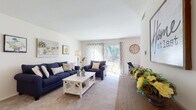 This single-floor residence offers a thoughtfully arranged layout that balances living, working, and resting spaces. The living room serves as the heart of the home, featuring plush wall-to-wall carpeting, neutral-toned walls, and generous natural light streaming through sliding glass doors that open to an outdoor view. The open sight line extends into a dedicated workspace area, complete with a chandelier overhead and additional windows dressed in patterned drapes, creating a bright and functional environment.

The kitchen is finished with warm oak cabinetry, tile flooring, and grey countertops. It includes a full suite of appliances—refrigerator, dishwasher, and range—alongside a stacked washer and dryer unit conveniently integrated into the space.

The bedrooms are carpeted and well-proportioned, each offering ample natural light through slatted blinds. The primary bedroom includes a large dresser with mirror, a seating area, and generous closet storage along one wall. A second bedroom provides a similarly comfortable and airy feel.

The bathroom features tile flooring, a wood-toned vanity with a wide countertop, a large mirror, and a full tub-shower combination, offering a clean and practical design throughout.