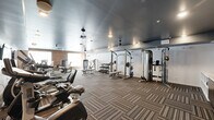 Located at 6715 Century Crest Pt in Colorado Springs, CO, this fitness center offers a well-appointed exercise room designed to support a comprehensive workout experience. The space is generously sized, providing ample room for a diverse selection of equipment and free movement throughout.

The cardio section features a row of treadmills and stationary bikes, including upright and recumbent models, all positioned beneath wall-mounted flat-screen TVs to keep users engaged during workouts. Ceiling fans are evenly distributed throughout the space, ensuring comfortable air circulation.

The strength-training area is equally well-equipped, housing a power rack, cable machines, and a full dumbbell rack with a variety of weight increments, accompanied by an adjustable weight bench. Additional selectorized machines provide targeted muscle group training options.

The flooring throughout consists of durable, patterned carpet tiles in neutral earth tones, adding a practical yet refined aesthetic. One wall is accented with a textured wood-look panel, lending warmth and visual interest to the interior. A built-in shelving unit and a stainless steel mini-refrigerator provide convenient storage and refreshment access, complemented by a dual-level water fountain nearby. Natural light filters in through windows, and recessed lighting ensures the space remains well-lit at all times.