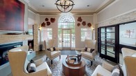 This well-appointed single-floor community space blends sophisticated design with functional social amenities. The grand entrance lobby sets an impressive tone with soaring ceilings, an arched transom window that floods the room with natural light, stacked stone accent columns, and an elegant orb chandelier. Wingback chairs arranged around a round wood coffee table create a refined yet inviting seating area, complemented by decorative wall art.

The lounge area transitions seamlessly into a comfortable sitting space featuring a leather sofa, patterned throw pillows, and bold typographic wall art that adds an eclectic, contemporary flair. Nearby, a game room offers a full-length shuffleboard table positioned beneath a large window with garden views, alongside vibrant red accent chairs adorned with decorative mirrors on soft grey walls.

The bar and entertainment area is a standout feature, showcasing a long white quartz-topped bar with modern saddle-style stools, stacked stone columns, a wood-paneled accent wall, dual wall-mounted televisions, a beverage refrigerator, and warm ambient wall sconces. Polished concrete flooring ties the social spaces together, lending an urban, contemporary edge to the overall design. This space is clearly designed for gathering, entertaining, and community engagement.