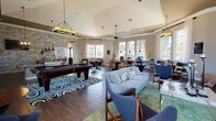 This single-floor clubhouse-style amenity space offers a well-appointed, open layout designed for both recreation and productivity. The main area is anchored by rich, dark wood-look tile flooring that flows throughout, complementing warm neutral-toned walls and elegant crown molding. Elevated ceilings with ceiling fans and stylish pendant lighting add dimension and comfort to the space.

A focal point of the room is a full-size billiards table set atop a bold, graphic area rug, surrounded by mid-century modern lounge seating including a plush sofa and accent chairs in soft grey and blue tones. Adjacent to the lounge, a dedicated business center lines the windowed perimeter, providing multiple computer workstations with ergonomic seating beneath tall, white-curtained windows that invite generous natural light and outdoor views.

A game and gathering area features round tables with mixed seating, ideal for board games or casual meetings. A stacked-stone accent wall adds a striking textural element near the kitchenette, which is outfitted with granite countertops, rich espresso cabinetry, a sink, and coffee-making appliances. Throughout, the space maintains a cohesive, polished ambiance suited for community use.