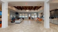 This single-floor community clubhouse is designed with an open-concept layout that seamlessly connects multiple functional zones under one roof. The entry foyer welcomes visitors with soaring ceilings, elegant herringbone-patterned tile flooring, and warm wood-framed glass doors that invite abundant natural light.

The main gathering area flows naturally between a lounge zone, game area, and a fully equipped kitchen. The lounge features a striking natural stone accent wall with a wall-mounted flat-screen TV, flanked by sconce lighting and accented by teal leather seating and vibrant orange side tables. Adjacent to the lounge, a billiard table sits beneath a coffered wood-beam ceiling with recessed lighting, adding a rustic-modern contrast to the otherwise contemporary design.

The kitchen is a standout feature, anchored by a curved marble-topped island with bar seating beneath a cascading cluster of globe pendant lights. Stainless steel appliances, a sleek range hood, and dark tile backsplash complement white cabinetry throughout. A dining area with modern chairs sits nearby, completing the entertaining space. Roller shades on large windows throughout the clubhouse provide light control while maintaining the bright, welcoming ambiance.