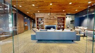 Located on Chapel Street in the heart of New Haven, CT, this thoughtfully designed single-floor commercial space blends industrial and warm organic aesthetics throughout. The entrance greets visitors with a dramatic floor-to-ceiling glass facade, flooding the interior with natural light and offering clear views of the surrounding streetscape. Polished terrazzo-style concrete floors extend seamlessly throughout, providing a refined yet durable foundation that ties the entire space together.

The welcoming reception area features a sleek natural wood desk set against cool grey walls, with warm reclaimed wood plank ceilings overhead — a design motif that carries consistently through the space. A wide corridor leads visitors deeper into the interior, where a spacious lounge area serves as the centerpiece of the space. Here, reclaimed wood wall paneling surrounds a modern fireplace flanked by open built-in shelving, creating an inviting and functional gathering area. Comfortable seating arrangements, including plush sofas and accent chairs, make the lounge ideal for collaboration or relaxation. Industrial-style pendant lighting complements the warm wood tones above. An adjacent area separated by glass partitions extends the usable space while maintaining an open, airy feel throughout.