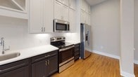 Located at 511 S Mangum St in the heart of Durham, NC, this single-floor unit offers a thoughtfully designed interior with contemporary finishes throughout. The open main living area is anchored by warm hardwood flooring that extends through the space, recessed lighting overhead, and a bright set of French doors that invite natural light and offer views of the surrounding urban streetscape.

The kitchen is a standout feature, showcasing a two-tone design with crisp white upper shaker cabinets and deep charcoal lower cabinetry, complemented by white quartz countertops and a classic subway tile backsplash. A full suite of stainless steel appliances — including a range, over-the-range microwave, refrigerator with ice maker, and dishwasher — completes this functional and stylish space. A decorative X-detail cabinet adds a refined architectural touch.

The bedroom is carpeted for comfort and features a large window with blinds that frames views of neighboring brick buildings, allowing for ample natural light. A barn-style sliding door adds a modern design element to the unit's interior.

The bathroom is well-appointed with dark espresso cabinetry, a dual-sink vanity with white countertops, a large mirror, and a walk-in glass-enclosed shower with white subway tile surround and chrome fixtures. The overall ambiance is clean, modern, and move-in ready.