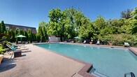 Located at 1314 Wharton Dr in Newark, Delaware, this outdoor recreational area centers around a generously sized swimming pool with clear, blue-green water. The pool is framed by a well-maintained brick-toned concrete deck that provides ample space for lounging, and is outfitted with multiple chaise lounge chairs and vibrant teal umbrellas, offering shaded seating for relaxation on sunny days. Large decorative terracotta planters are positioned throughout the deck, adding a touch of greenery and visual warmth to the setting. The entire pool area is enclosed by a sturdy black wrought iron fence, providing both security and an unobstructed view of the surrounding landscape. Mature trees create a natural, private canopy that lines the perimeter, lending a sense of seclusion and tranquility. A utility shed is conveniently situated within the enclosure. Adjacent to the pool, a grassy lawn area offers additional open space, and a playground is visible nearby, making this an amenity suited for a range of ages. The overall ambiance is bright, welcoming, and well-maintained, presenting an ideal outdoor retreat during warm months beneath open blue skies.