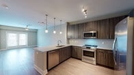 Located at 9200 Bruckhaus St in Raleigh, NC, this single-floor residence offers a thoughtfully designed open-concept layout with contemporary finishes throughout. The kitchen serves as a central highlight, featuring rich dark wood cabinetry with brushed gold hardware, stone countertops, a subway tile backsplash, and a full suite of stainless steel appliances including a side-by-side refrigerator, electric range, built-in microwave, and dishwasher. A peninsula with an integrated sink extends into the living area, ideal for casual dining or entertaining. Pendant lighting and track fixtures add warm illumination throughout the kitchen and open living space. The living area is finished with durable wood-look flooring and is anchored by French doors that invite natural light and provide outdoor access. A ceiling fan enhances comfort in both the living and bedroom spaces. The carpeted bedrooms offer a cozy retreat, each with natural light from window blinds and organized closet space with built-in shelving and hanging rods. The bathroom features a soaking tub with a full subway tile surround and brushed nickel fixtures, providing a clean and polished aesthetic. The overall design balances modern style with practical functionality.