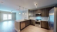 Located at 9200 Bruckhaus St in Raleigh, NC, this single-floor residence offers a thoughtfully designed open-concept layout with contemporary finishes throughout. The kitchen serves as a central highlight, featuring rich dark wood cabinetry with brushed gold hardware, stone countertops, a subway tile backsplash, and a full suite of stainless steel appliances including a side-by-side refrigerator, electric range, built-in microwave, and dishwasher. A peninsula with an integrated sink extends into the living area, ideal for casual dining or entertaining. Pendant lighting and track fixtures add warm illumination throughout the kitchen and open living space. The living area is finished with durable wood-look flooring and is anchored by French doors that invite natural light and provide outdoor access. A ceiling fan enhances comfort in both the living and bedroom spaces. The carpeted bedrooms offer a cozy retreat, each with natural light from window blinds and organized closet space with built-in shelving and hanging rods. The bathroom features a soaking tub with a full subway tile surround and brushed nickel fixtures, providing a clean and polished aesthetic. The overall design balances modern style with practical functionality.