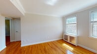 This single-floor residence presents a well-maintained layout with consistent design elements throughout. Warm-toned hardwood floors run through every living area, reflecting natural light that streams in generously through multiple windows. Crown molding adds a classic architectural detail to several rooms, lending a refined character to the interiors.

The living spaces are bright and open, with neutral wall tones creating a clean, versatile backdrop. Several rooms are equipped with ceiling fans and traditional radiator heating units positioned beneath the windows, indicating a well-established heating system. Each room benefits from one or more windows, offering views of the surrounding neighborhood and greenery.

The kitchen features white cabinetry with raised-panel detailing, dark granite-style countertops, a mosaic tile backsplash, recessed lighting, and a black gas range. A large window overlooks outdoor greenery, adding brightness to the cooking space. A peninsula counter provides a natural division between the kitchen and adjacent areas.

The bathroom retains a classic character, showcasing original black-and-white basketweave tile flooring, white subway-style wall tiles with black accent trim, and a tub-shower combination. A window above the tub brings in natural light, enhancing the room's functionality and feel.

Overall, the space balances traditional architectural details with practical, updated features throughout.