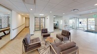 This single-floor amenity space is designed with a cohesive modern aesthetic, characterized by light herringbone-patterned hardwood flooring, crisp white walls, and an abundance of natural light streaming through large floor-to-ceiling windows. The main lounge area makes a striking first impression with a sculptural gold sputnik chandelier suspended from a tray ceiling, complemented by a plush sectional sofa, a marble-topped coffee table, and curated wall art. Glass-walled partitions throughout the space create a sense of openness while defining distinct functional zones. A seating lounge features sleek chrome-framed upholstered chairs arranged around organic wood accent tables, with views into adjacent glass-enclosed rooms. A game room, visible through full-height sliding glass doors, includes a foosball table and pool table set against patterned accent walls. A communal kitchen and dining area offers bar-height seating and pendant lighting. The entry corridor features a state-of-the-art automated parcel locker system alongside wood-tread stairs. A glass-enclosed fitness center rounds out the amenities. The overall ambiance is polished, bright, and inviting.