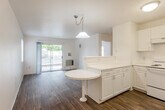 Located at 1725 Almaden Road in San Jose, CA, this single-floor, one-bedroom, one-bathroom residence offers a compact yet efficient layout with wood-look flooring throughout. The open-concept living and dining area benefits from natural light streaming in through a sliding glass door that leads to an outdoor space, creating an inviting and airy atmosphere. A pendant light fixture anchors the dining area, which features a built-in round table extending from the kitchen peninsula — a practical and space-conscious design detail. The kitchen is equipped with white cabinetry, a stainless steel refrigerator, a white electric range, and light speckled countertops that extend along the peninsula, providing ample prep and storage space. The bedroom is a comfortable, well-proportioned room featuring two windows with blinds that allow for natural light and privacy, a ceiling fan with overhead light, and a dedicated closet accessible through a paneled door. The bathroom is thoughtfully designed with accessibility features, including grab bars, a wall-mounted sink, white subway tile accents along the lower walls, and wood-look flooring consistent with the rest of the unit. Clean white walls and consistent finishes throughout lend the space a bright, cohesive, and move-in ready character.